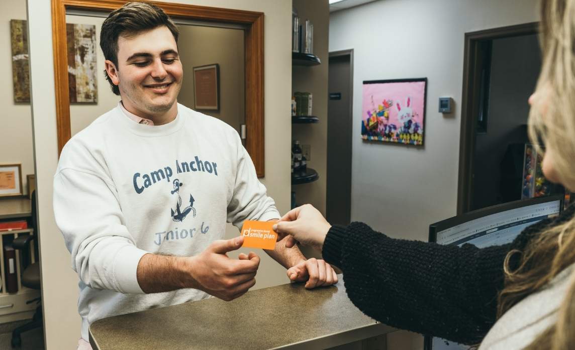pd-smileplan card A smiling young man accepts an orange Progressive Dental Smile Plan card from a patient a staff member at the front desk.